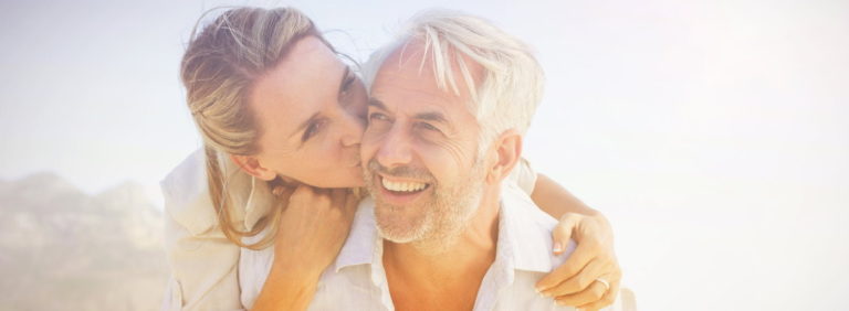 Middle aged couple smiling while the women kisses the man on the cheek in the sunlight.