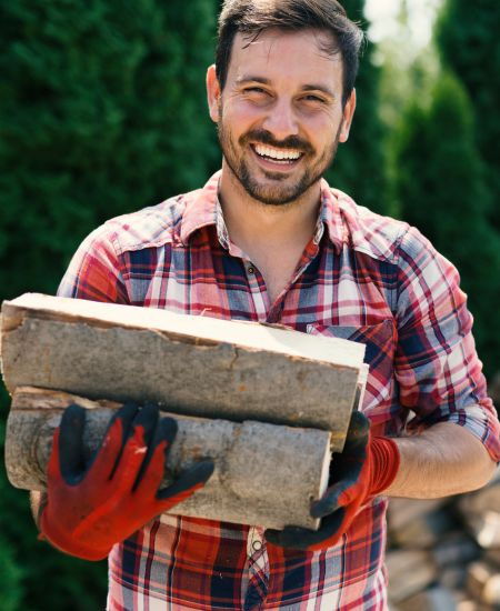 A young man in a flannel shirt smiles at the camera while carrying firewood.
