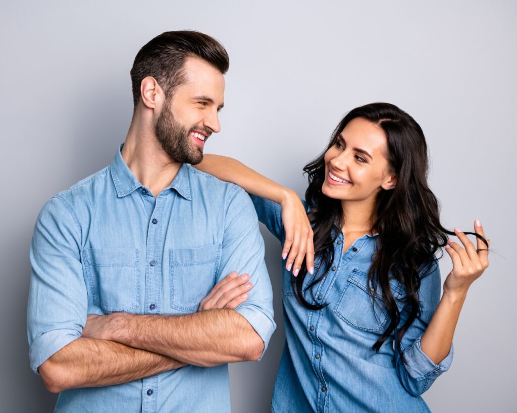 A couple with dark hair and blue shirts look at each other and smile on a grey background.