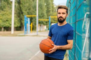 A man with brown hair and a leans against a fence on a basketball court while holding a basketball and looking at the camera